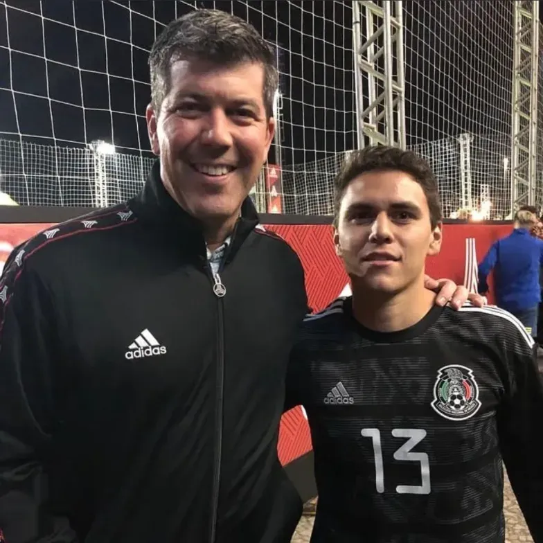 Dos personas en una cancha por la noche, a la derecha una persona con una camiseta del equipo de selección mexicana, a la izquierda Fernando Palomo