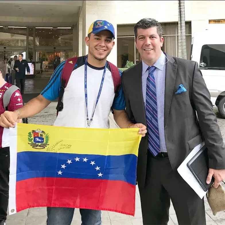 Fuera de un edificio, a la izquierda persona sosteniendo una bandera venezolana y a la derecha Fernando Palomo